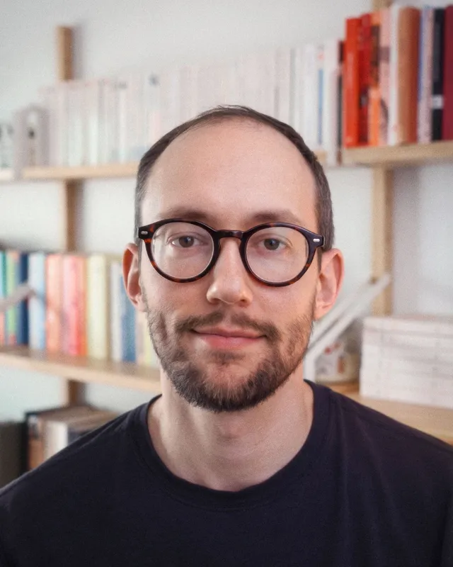 Portrait de Didier Guyaux, lunettes rondes en écaille, devant une bibliothèque.